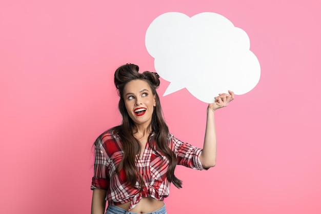 Young pinup woman in retro outfit holding blank white speech bubble on pink studio background