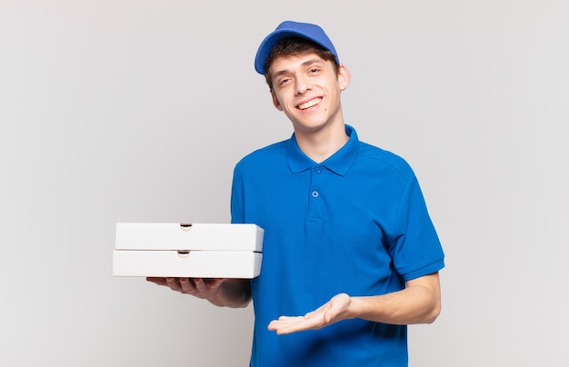 Young pizza deliver boy smiling cheerfully, feeling happy and showing a concept in copy space with palm of hand