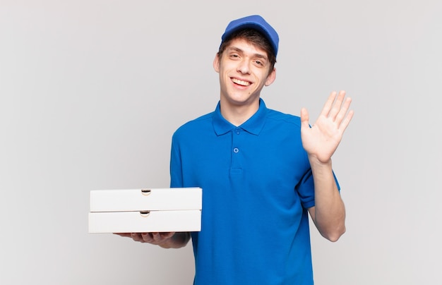 Молодой мальчик-доставщик пиццы радостно и весело улыбается, машет рукой, приветствует и приветствует вас или прощается