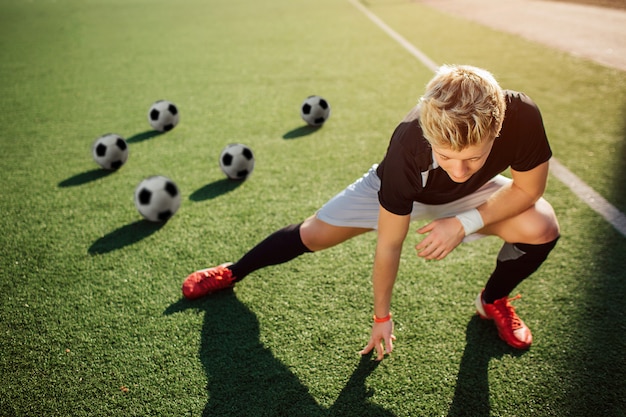 Photo young player stretch on green lawn. he does exercises for legs. guy look down. four balls are behind him.