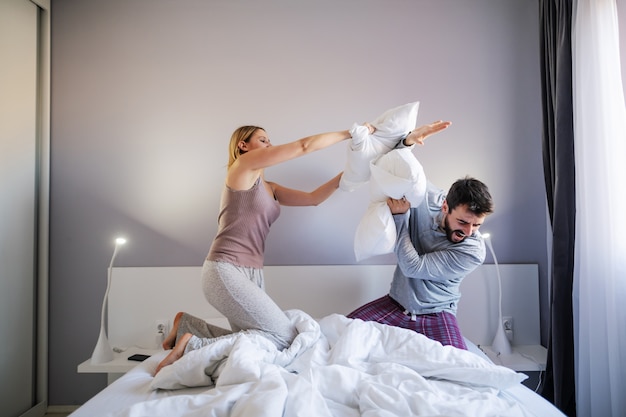 Young playful couple having pillow fight in the morning in bedroom