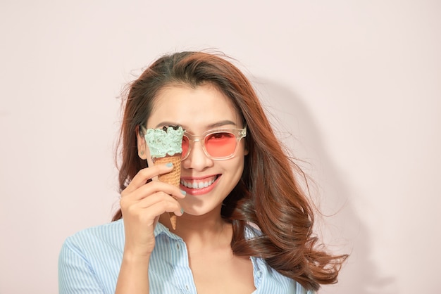 Young playful in eyeglasses covering eye with ice-cream cone on light background.