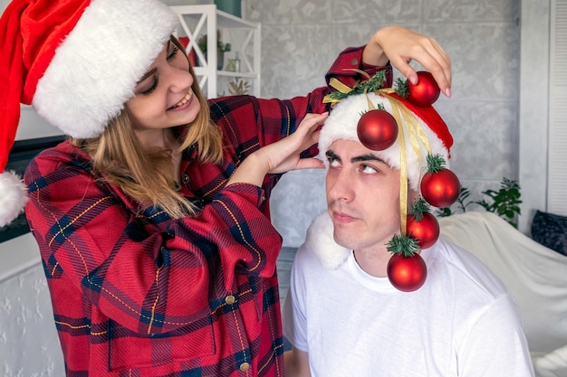 Foto giovane donna allegra che scherza addosso a decorare l'uomo con le palle di natale invece dell'albero di natale uomo che la guarda con rabbia coppia che indossa cappelli di babbo natale e pigiama rosso