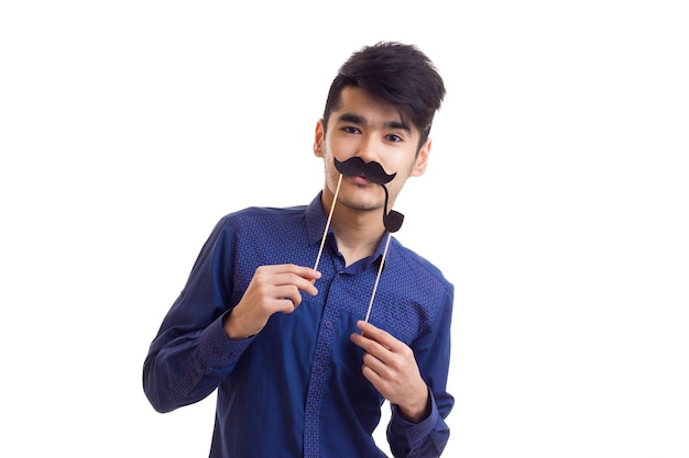 Young pleasant man with dark hair in blue shirt holding cardboard stick of moustache in studio