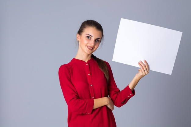 Young pleasant woman in blouse with plait holding white sheet of paper on grey background in studio