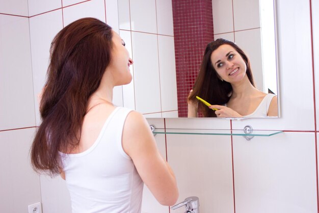 Young pleasant woman brushing her long brown hair with comb in front of the mirror in bathroom