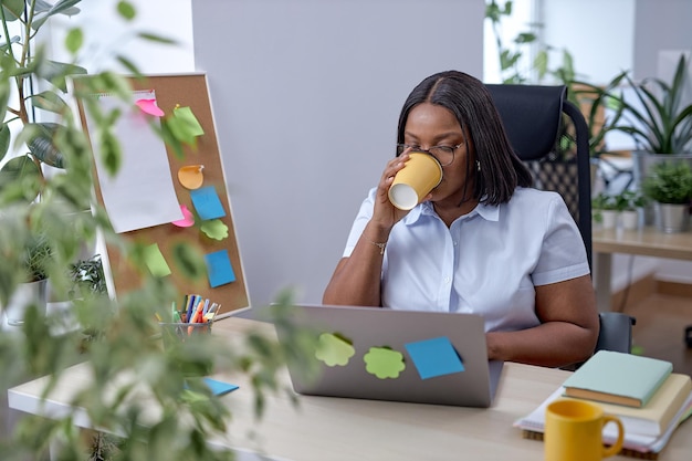 若い楽しい女性はお茶を飲みながらオフィスの机に座って、職場で休憩します。