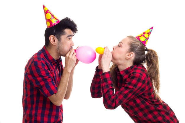 Giovane donna piacevole con una lunga coda di cavallo castagna e un giovane bell'uomo in camicie a quadri rosse con cappelli celebrativi che soffiano palloncini su sfondo bianco in studio