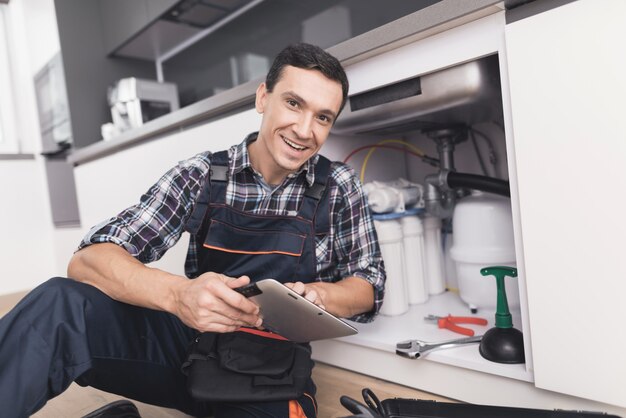 Young Plumber Is Sitting Near The Sink With A Tablet.