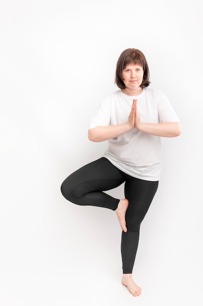 Young plump woman practices yoga. Portrait of girl in tree pose on white background.