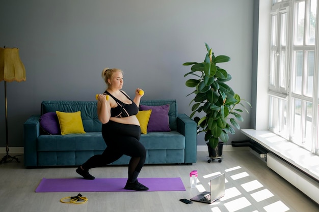 Young plump woman trains with yellow dumbbells lunging and\
squatting forward