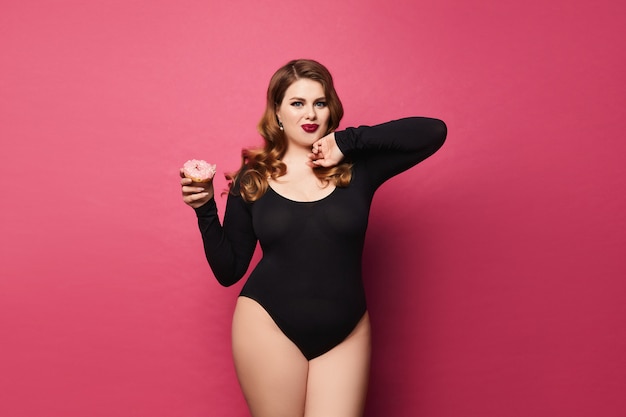Young plump woman with bright makeup in black bodysuit is holding dessert in her hand and posing at pink background, isolated with copy space.