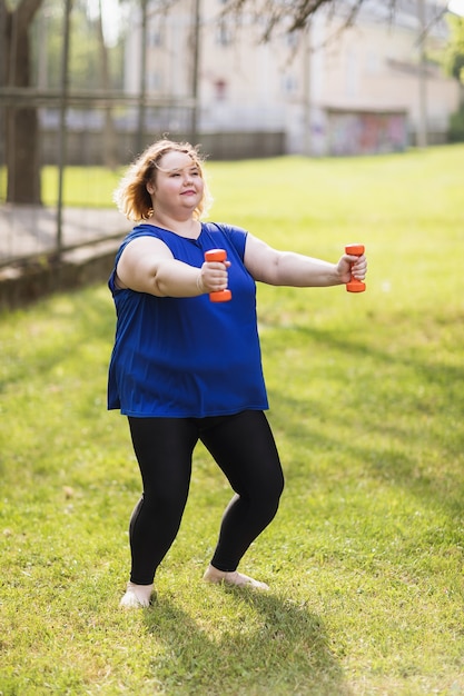 A young plus size blonde performs an exercise with dumbbells outdoors