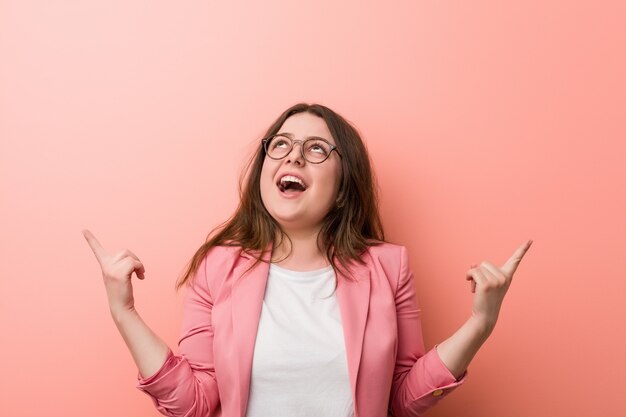 Young plus size business caucasian woman indicates with both fore fingers up showing a blank space.