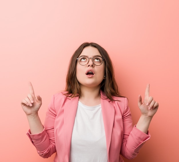 Young plus size business caucasian woman pointing upside with opened mouth.