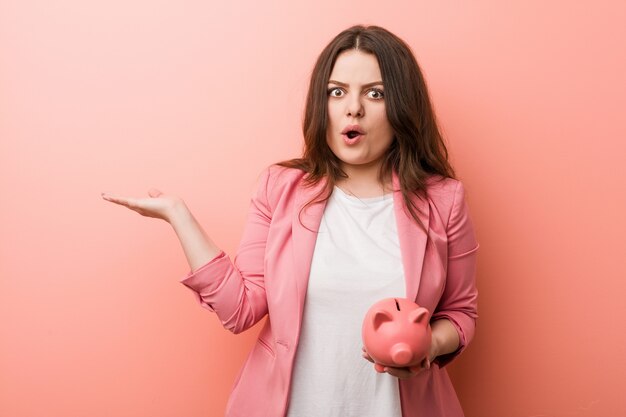 Young plus size curvy business woman holding a piggy bank impressed holding copy space on palm.