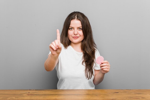 Young plus size curvy woman holding a facial sponge showing number one with finger.