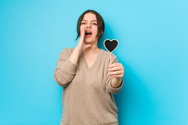 Young plus size curvy woman holding a heart shape shouting excited to front.