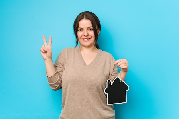 Young plus size curvy woman holding a home icon showing number two with fingers.