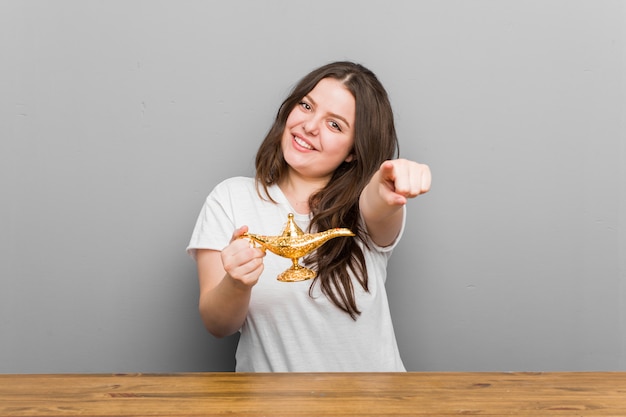 Young plus size curvy woman holding a magic lamp cheerful smiles pointing to front.