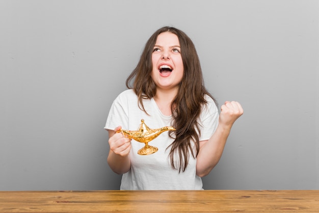 Young plus size curvy woman holding a magic lamp cheering carefree and excited. Victory concept.