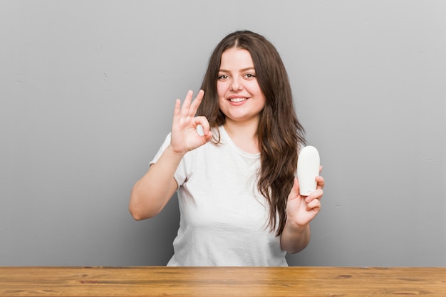 Young plus size curvy woman holding a moisturizer cheerful and confident showing ok gesture.