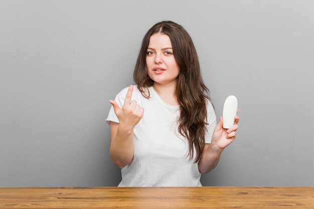 Young plus size curvy woman holding a moisturizer pointing with finger at you as if inviting come closer.