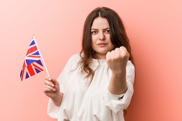 공격적인 표정으로 주먹을 보여주는 영국 국기를 들고 젊은 더하기 크기 매력적인 여자.