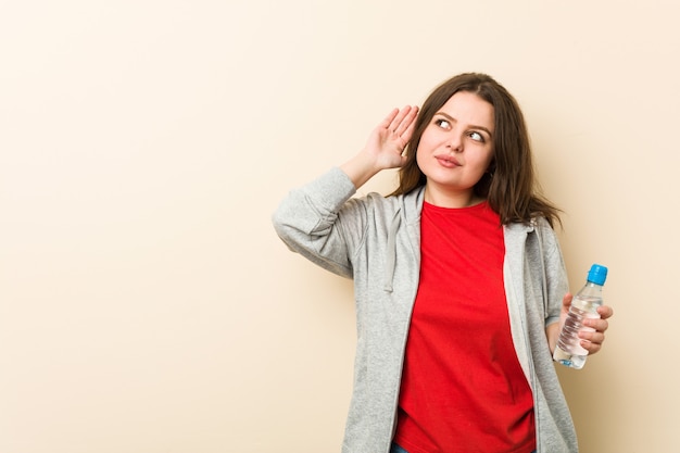 Young plus size curvy woman holding a water bottle trying to listening a gossip.