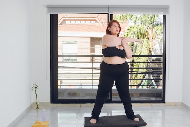 Young plus size latin woman in black sportswear doing arm stretch indoors