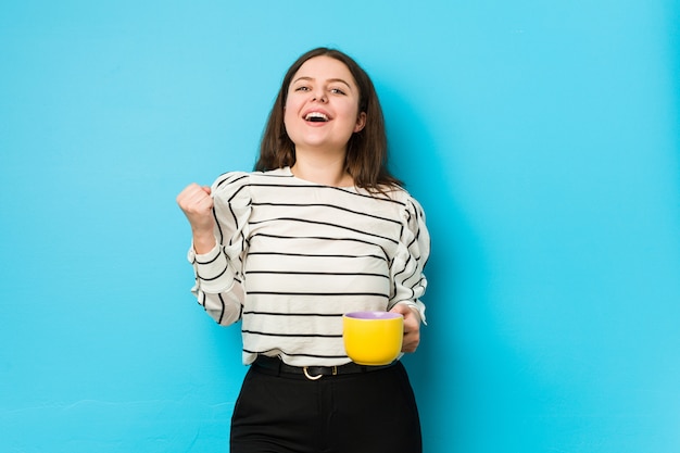 Young plus size vrouw met een theemok zorgeloos juichen en opgewonden. overwinning concept.