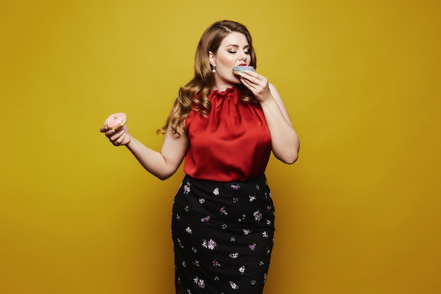 Young plus size woman, fashionable blonde girl with bright makeup, in red satin blouse and black skirt, with donuts in her arm posing with closed eyes