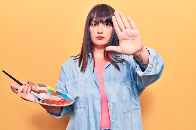 Young plus size woman holding paintbrush and palette with open hand doing stop sign with serious and confident expression, defense gesture