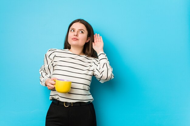 Young plus size woman holding a tea mug trying to listening a gossip.