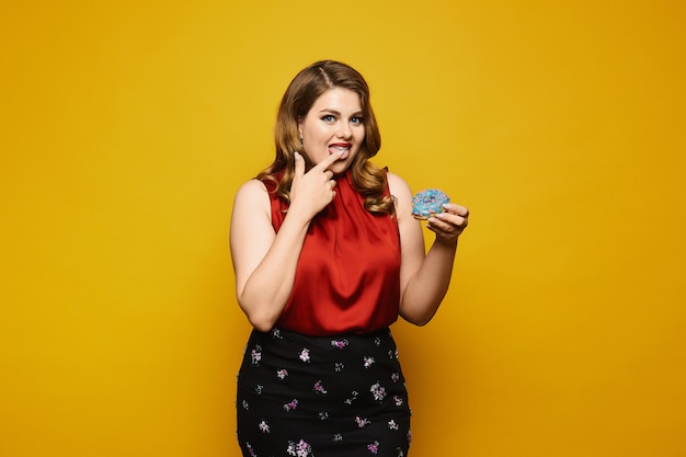 Young plus size woman in red blouse and skirt eating sweet donut on yellow , isolated