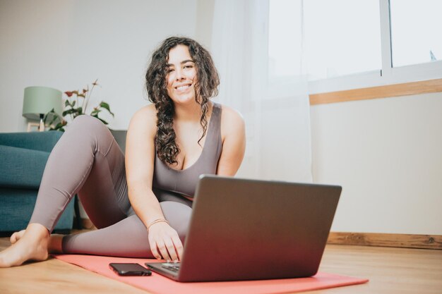 Young plus size woman sitting on the floor resting after doing a online yoga class with his laptop. Smiling to camera and happy about loosing weight with courses. Sporty clothes.