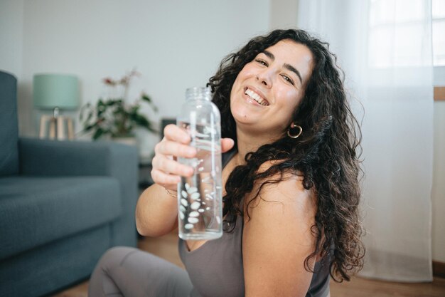 Giovane donna più grande che sorride all'acqua potabile della telecamera mentre fa fitness e yoga a casa seduto sul tappetino da yoga per pavimento. perdere peso a casa dopo le vacanze. capelli ricci lunghi.