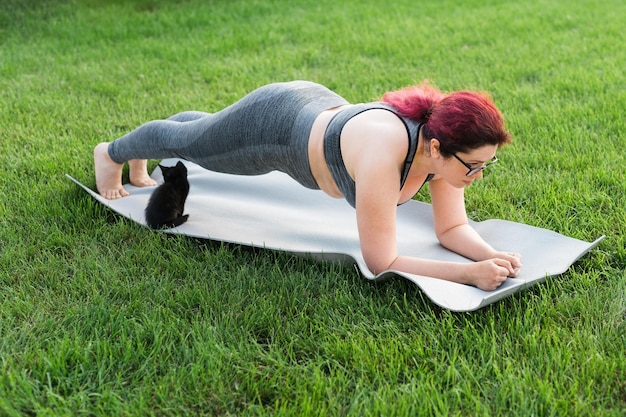 Young plus size woman in sporty top and leggings standing in plank on yoga mat spending time on green grass in yard Black kitten walks around her Well being and body positivity fitness concept