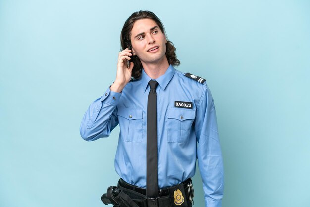 Young police caucasian man isolated on blue background keeping\
a conversation with the mobile phone with someone