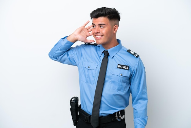 Young police caucasian man isolated on white background listening to something by putting hand on the ear