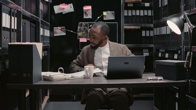 Young policeman examining forensic evidence in office, looking at case files with criminal records. Law enforcement officer reading reports and suspicious statements. Handheld shot.