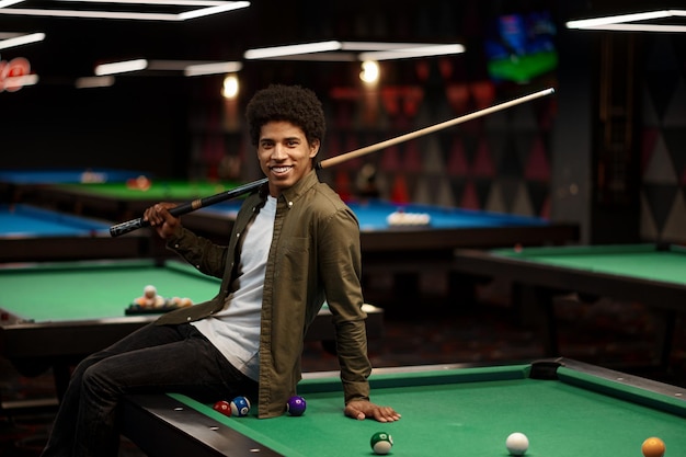 Young pool player with cue sitting on billiard table