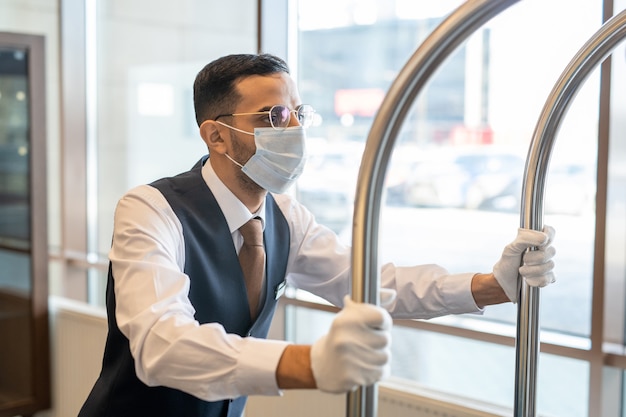 Young porter in uniform, eyeglasses and protective mask pushing cart with baggage while moving along corridor inside contemporary hotel