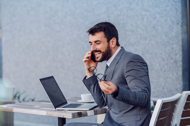 Giovane uomo d'affari caucasico positivo in vestito che si siede al tavolo, tenendo gli occhiali e avendo telefonata con sua moglie. sul tavolo ci sono laptop e caffè.