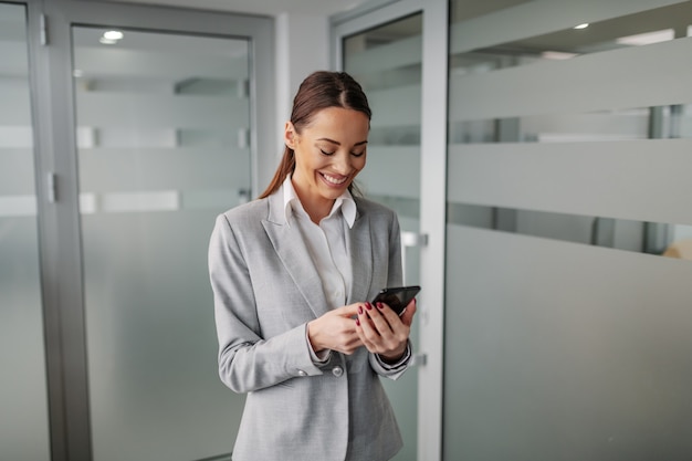 Young positive caucasian businesswoman in suit standing inside of corporate firm and using smart phone for reading an e-mail.