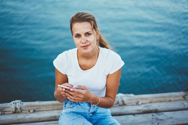 Foto giovane donna caucasica positiva che guarda la fotocamera durante la prenotazione dell'hotel per il prossimo viaggio tramite telefono cellulare