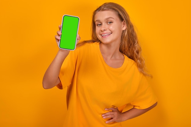 Young positive caucasian woman shows phone with green screen stands in studio