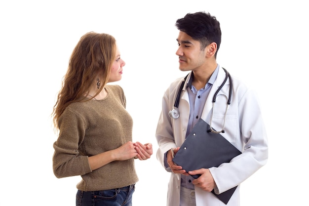 Young positive doctorin white gown holding a folder and talking to young pretty woman in sweater