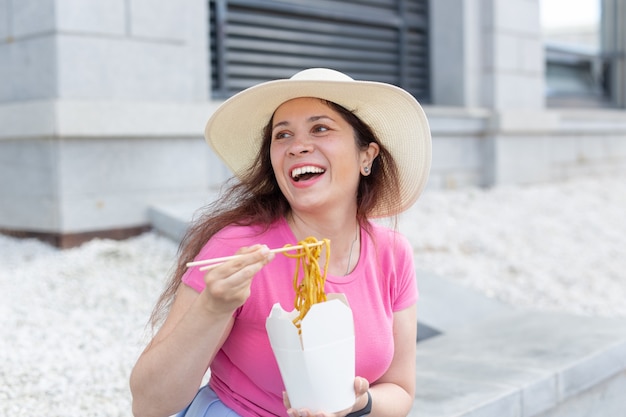 Young positive girl in a hat eats chinese noodles with wooden chopsticks for take-away lunch. Concept of high-calorie healthy food.