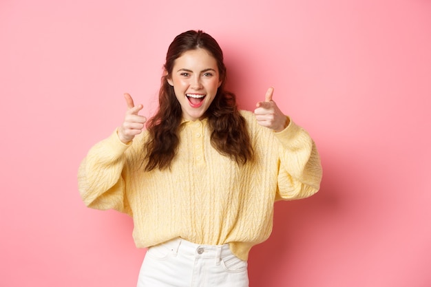 Young positive girl looking at you with confident and pointing camera inviting customers check out promo offer standing upbeat and smiling against pink wall